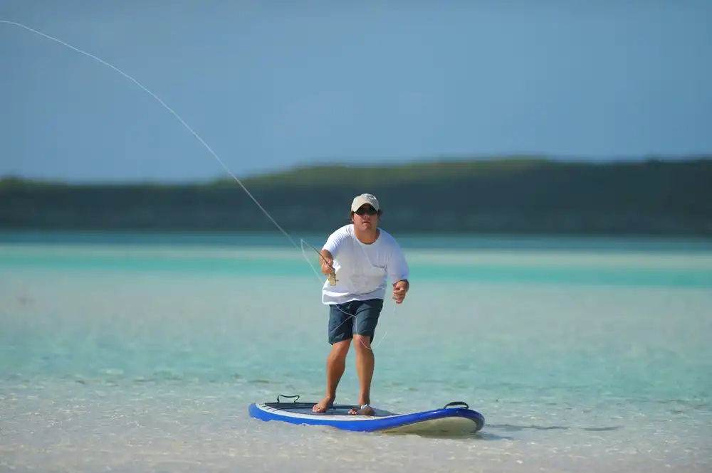 Muž rybařící na paddleboardu v mělké tyrkysové vodě, ukázka paddleboardu na rybaření.